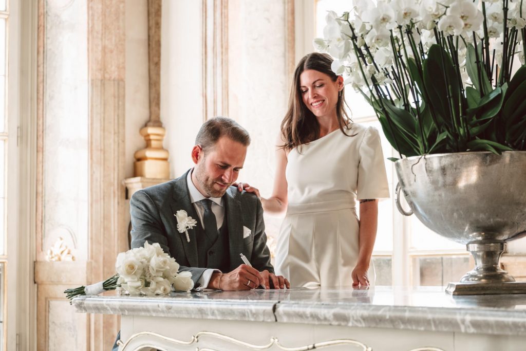 Die Eheurkunde wird unterschrieben. Sie steht freudig hinter ihm.